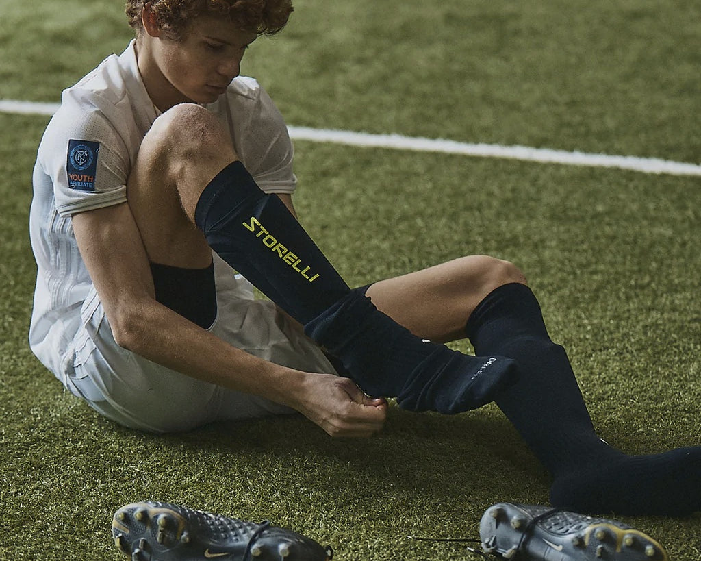 A young player adjusts his leg guards on a soccer field. 