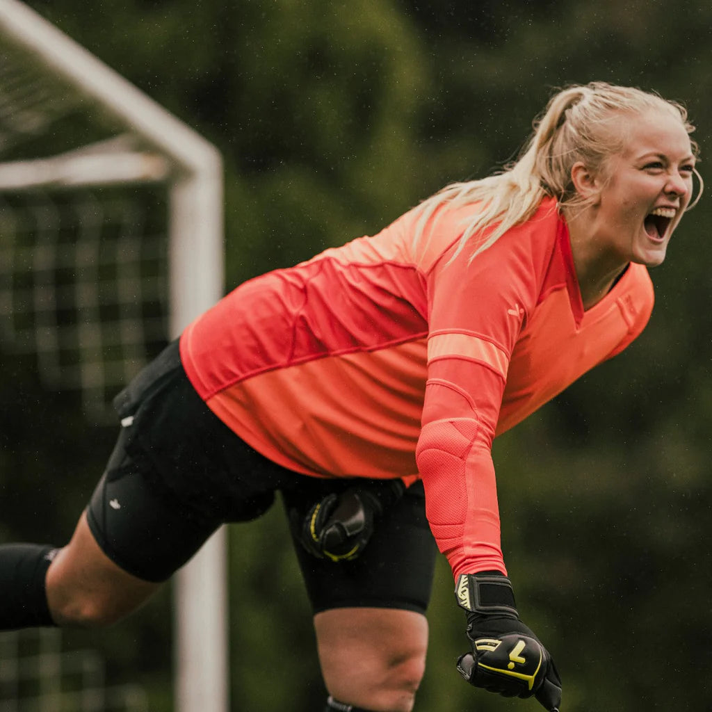 A female goalkeeper leans forward on foot, shouting in celebration. 
