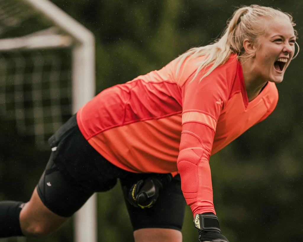 A female goalkeeper leans forward on foot, shouting in celebration. 